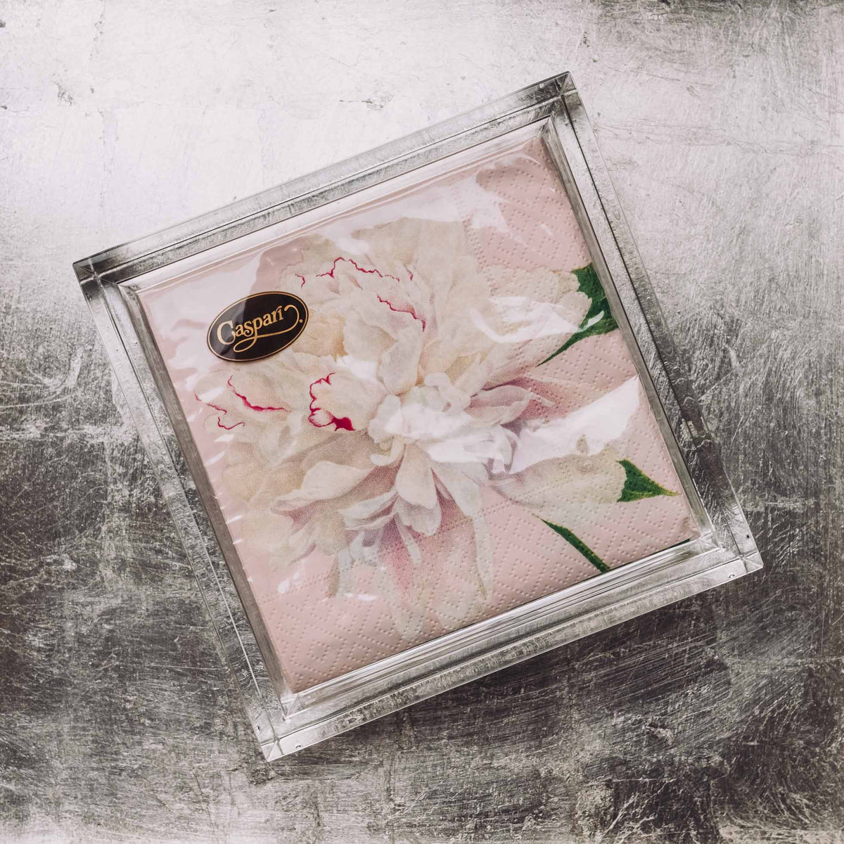 Clear acrylic napkin holder with gold trim containing pink and white peony-print napkins with green leaves