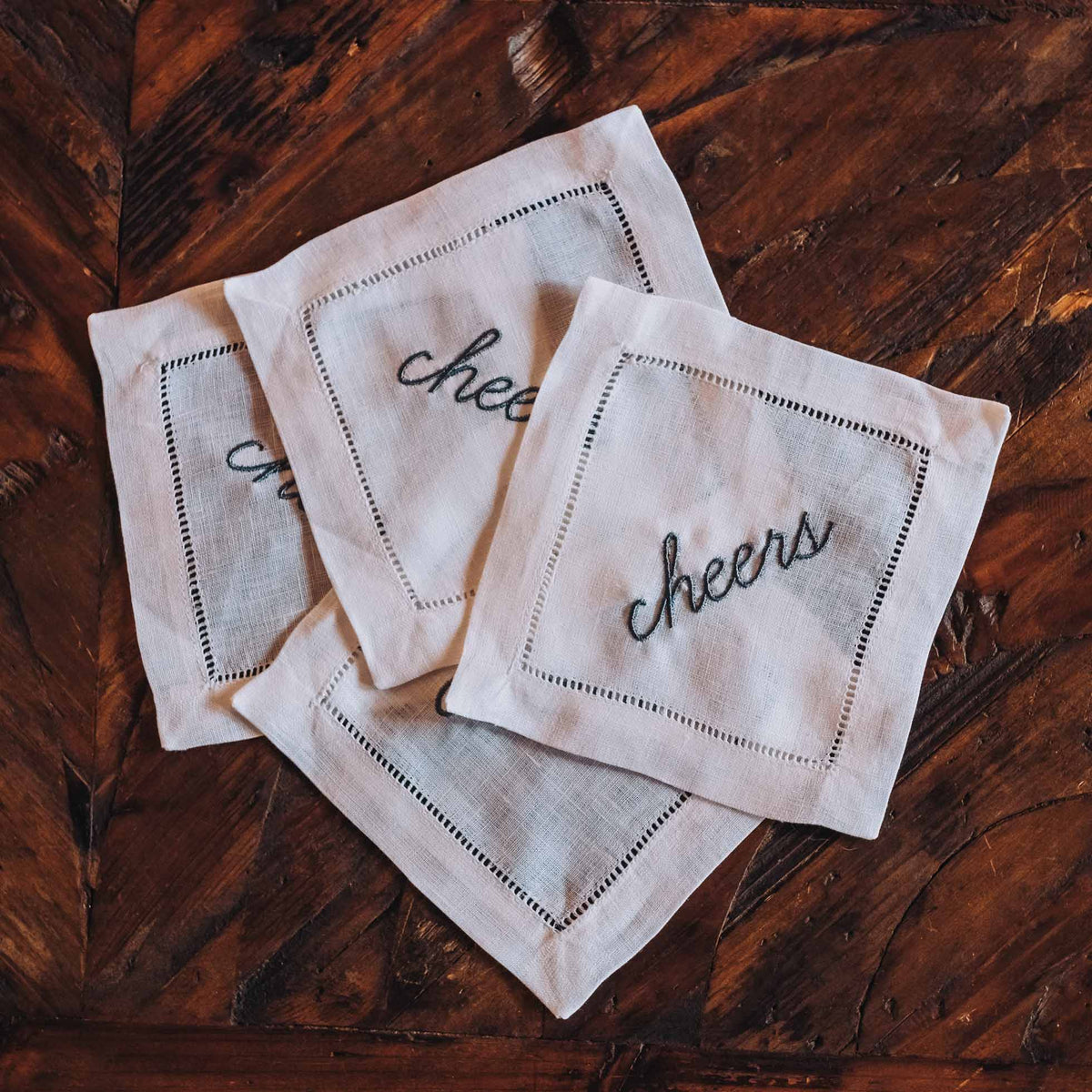 Set of four linen coasters with charcoal grey "cheers" embroidery and whipped edges on dark wood surface.