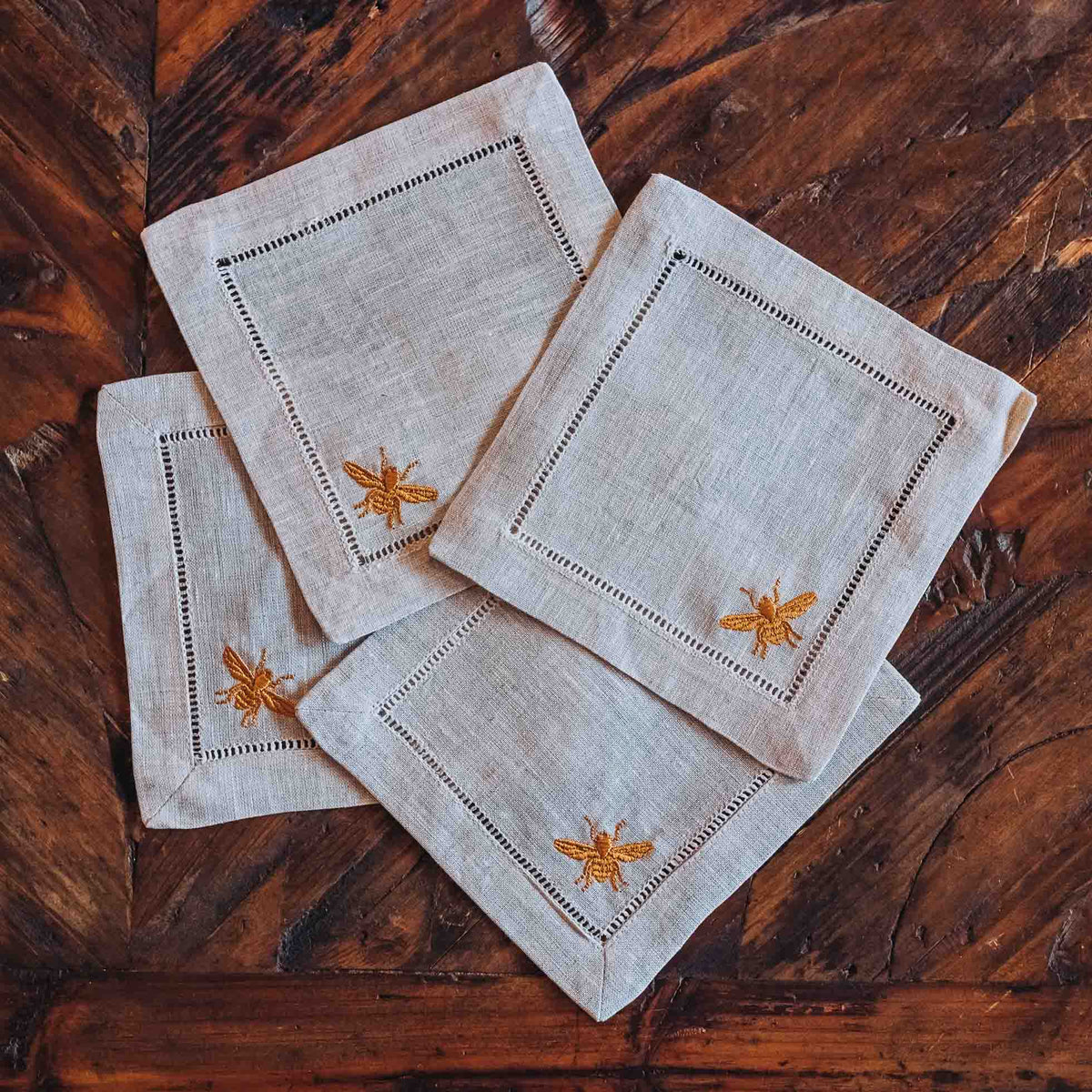 Set of three square linen cocktail coasters with hemstitched edges and gold embroidered bee motifs on natural grey linen, arranged on wood surface.