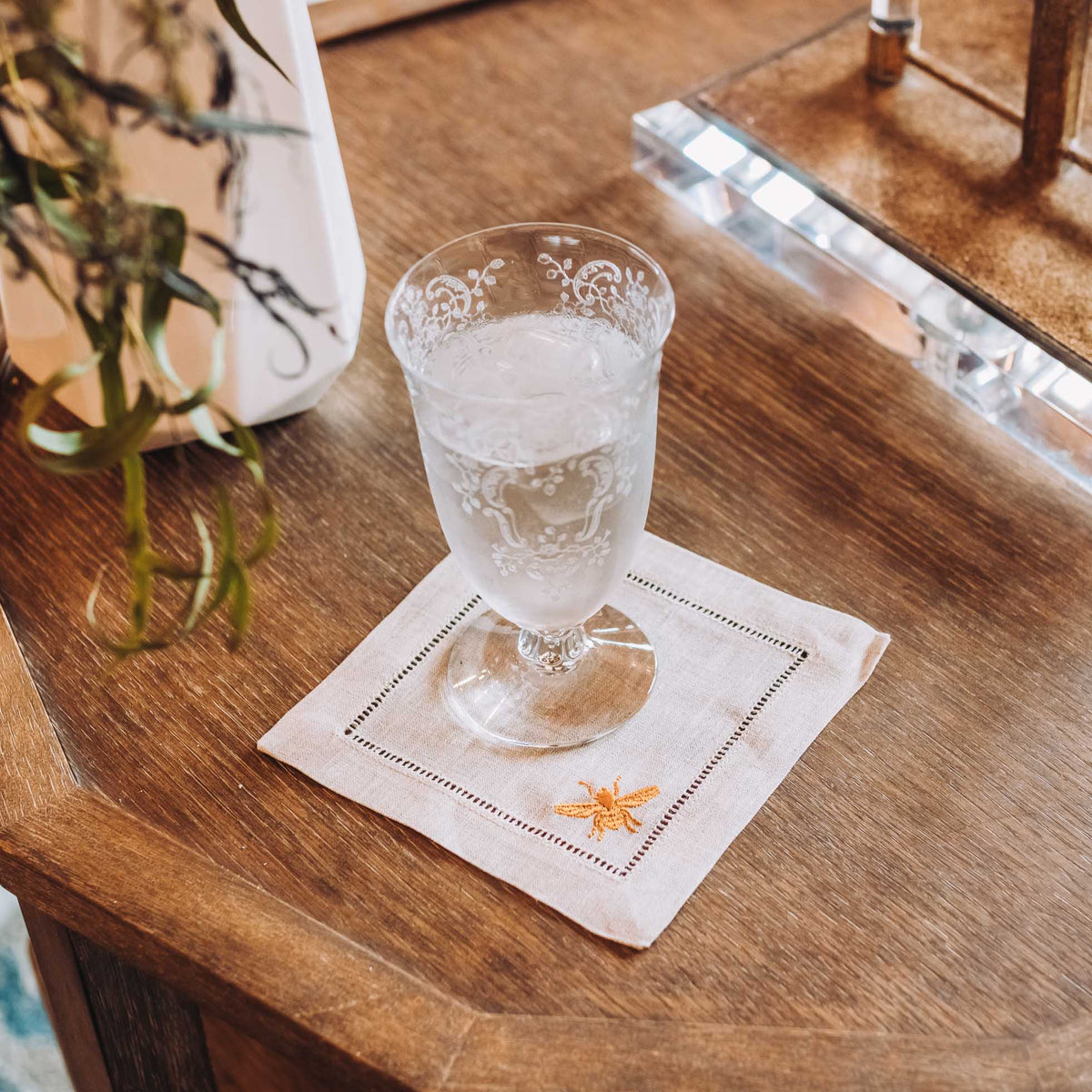 Square linen coaster with embroidered bee design in coral and aqua, placed under a cold drink on wooden surface