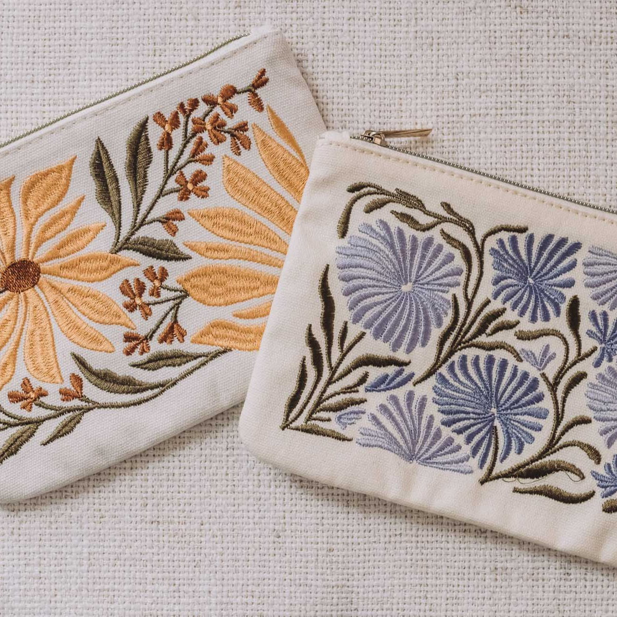 Two cream zip pouches with embroidered flowers: yellow sunflower and purple allium designs with green stems on natural linen fabric