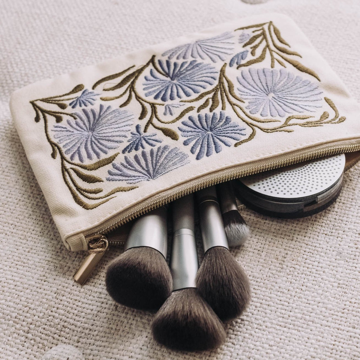 Cream zip pouch with embroidered blue and gold allium and sunflower flowers, featuring gold metal zipper