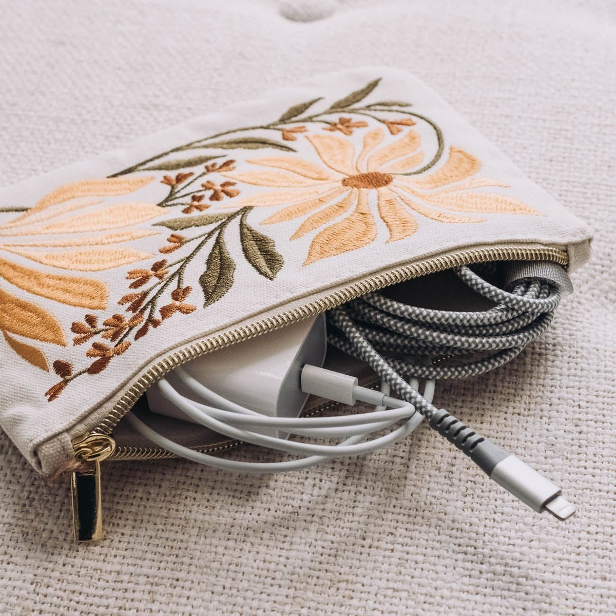 Cream zip pouch embroidered with yellow sunflowers and olive allium stems, open to reveal coiled cables inside