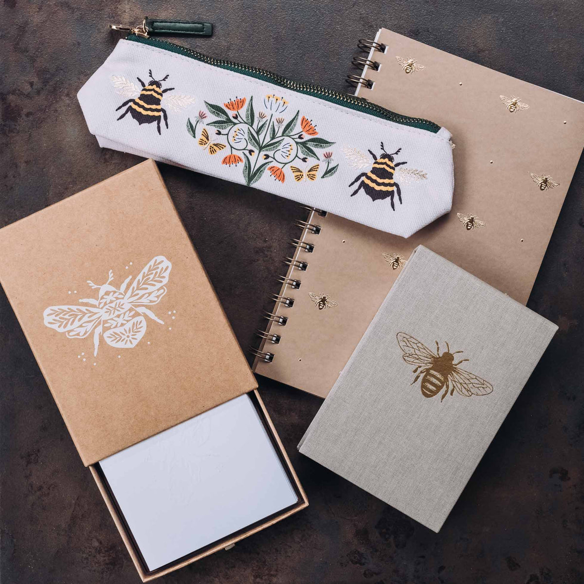 Collection of bumble bee stationery including a white pencil case, spiral notebooks, and notecards with embossed and illustrated bee designs on neutral kraft and linen backgrounds
