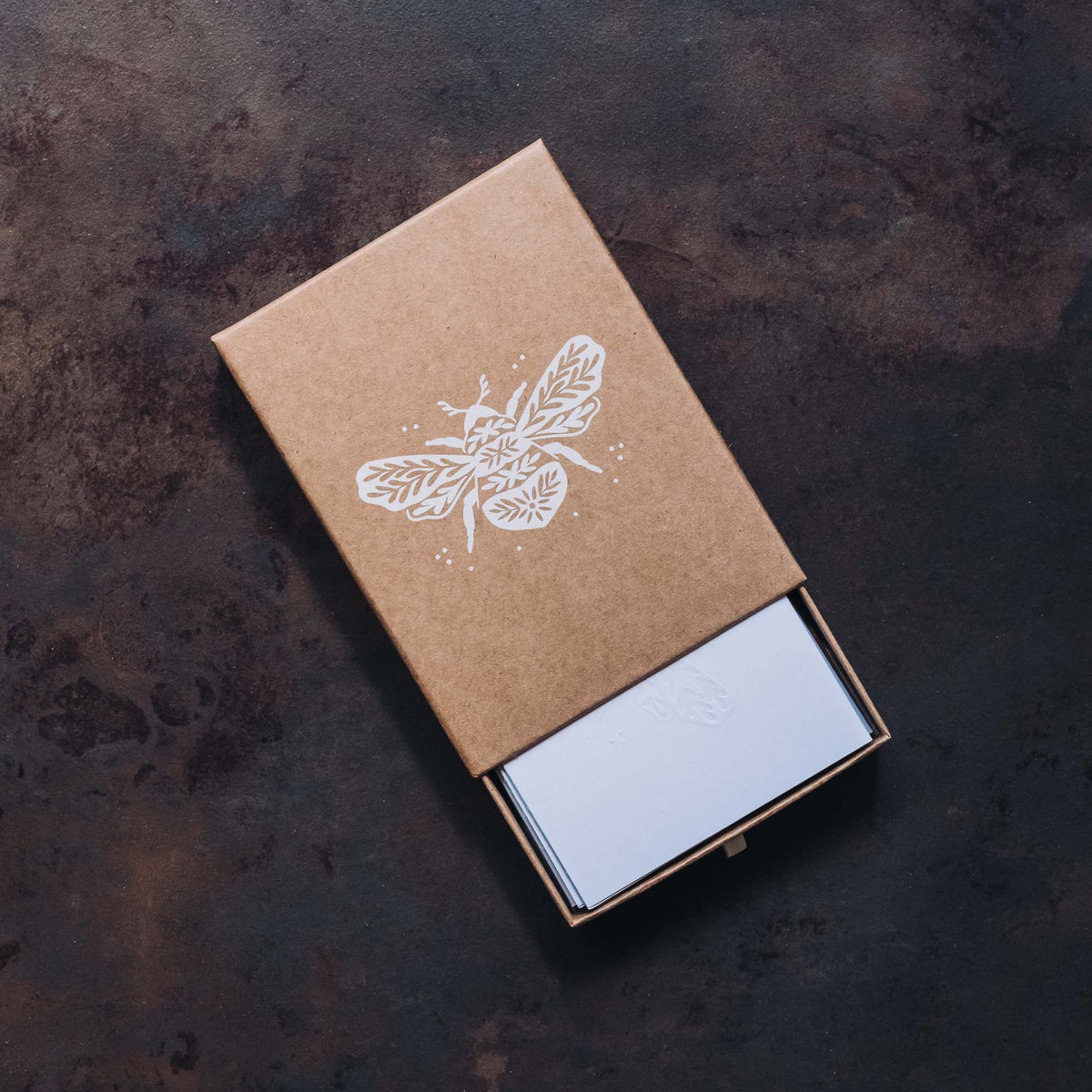 Kraft brown notecard box with white embossed bumble bee illustration, partially open to show white notecards inside