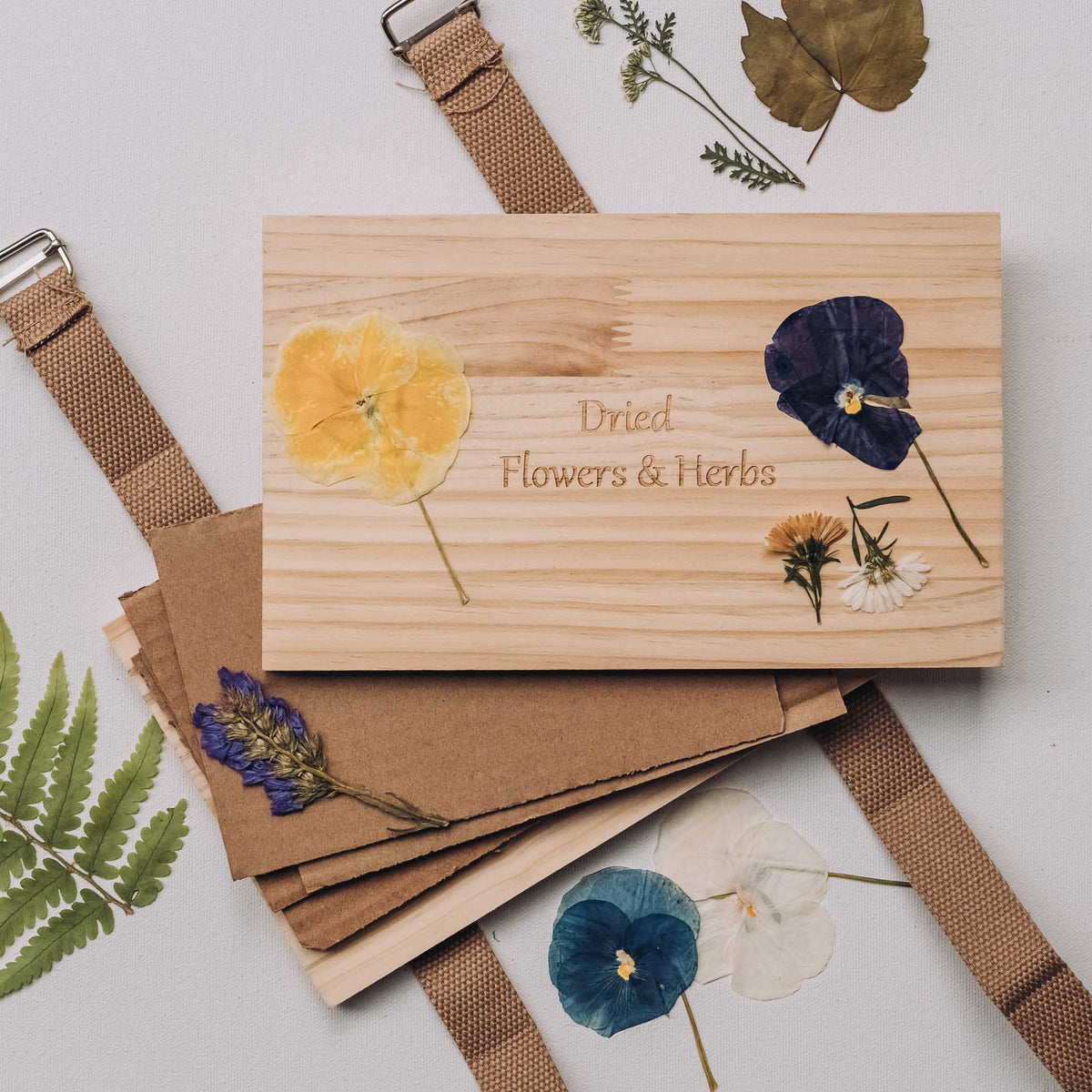 Wooden flower and herb press with pressed yellow and navy blue flowers engraved on light wood surface, secured with tan fabric straps.