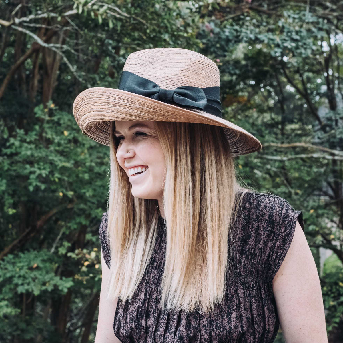 Woman wearing hand-woven straw hat with navy ribbon band and asymmetrical brim, styled outdoors against lush green foliage