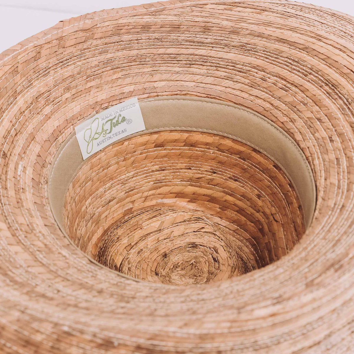 Hand-woven palm straw hat with asymmetrical brim in natural tan, showing UPF 50+ sun protection with woven texture detail and Tula brand label.
