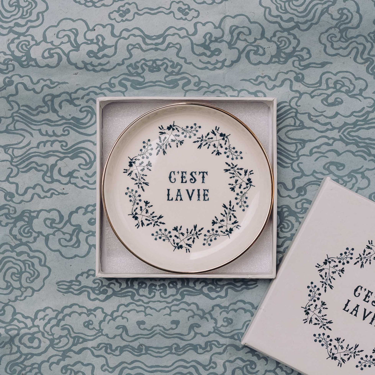 Cream ceramic trinket dish with "C'est La Vie" text and navy leaf wreath design, displayed in white box insert