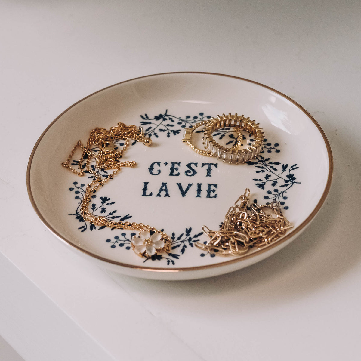 Round ceramic dish with cream glaze, navy florals, "C'est La Vie" text, and rose gold rim, styled with gold chain necklaces and ornate bangles.