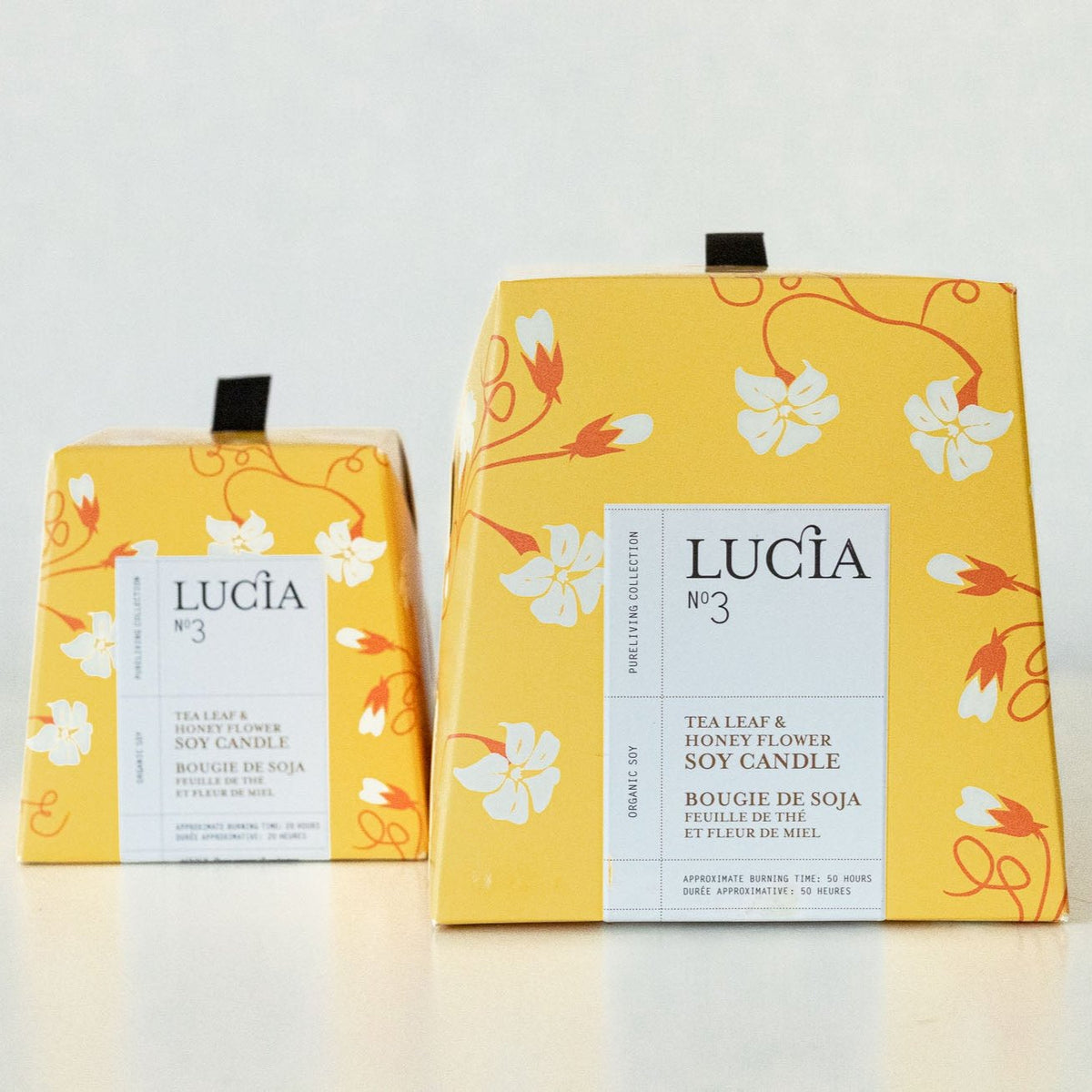Two Lucia No. 3 soy candles in bright yellow packaging with red scrollwork and white florals, votive and larger sizes side by side on cream surface.
