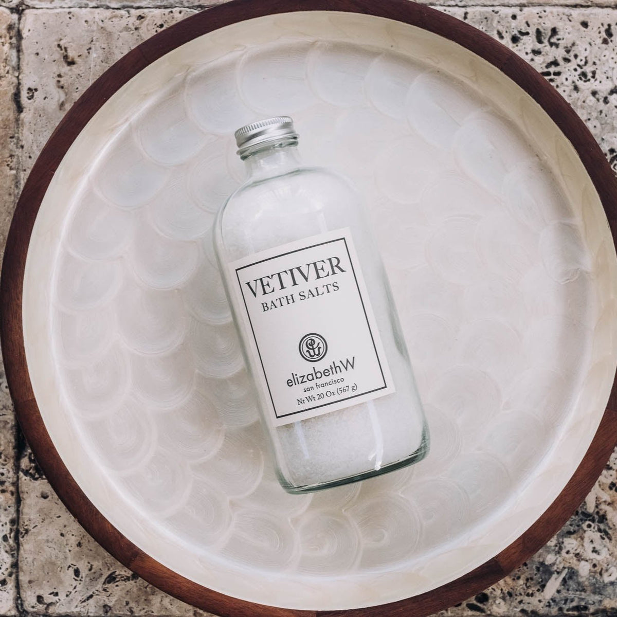 Vetiver bath salts in a clear glass bottle with metal cap, resting in a ceramic dish on a terrazzo surface