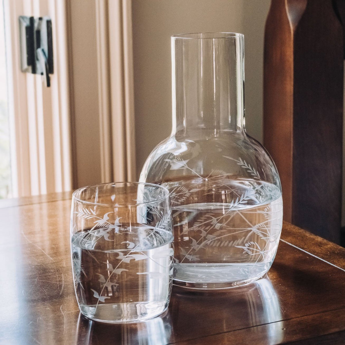Clear crystal carafe and matching glass with hand-engraved fern design, displayed on wood table