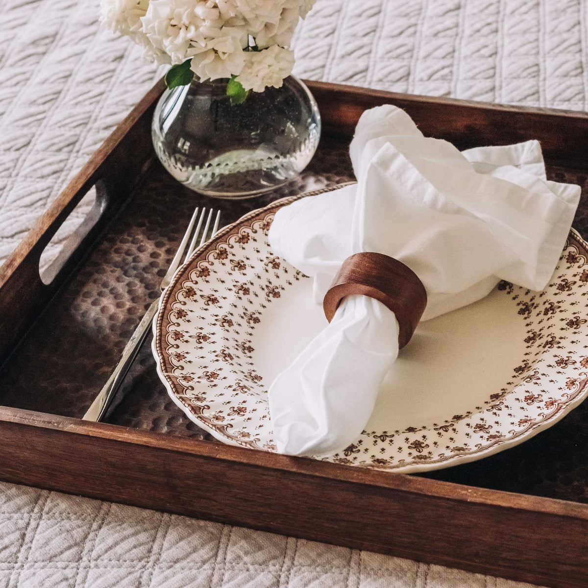 Brown hammered wood serving tray with inset handles, displaying elegant place setting with patterned plates, white napkin, and ivory flowers in glass vase.