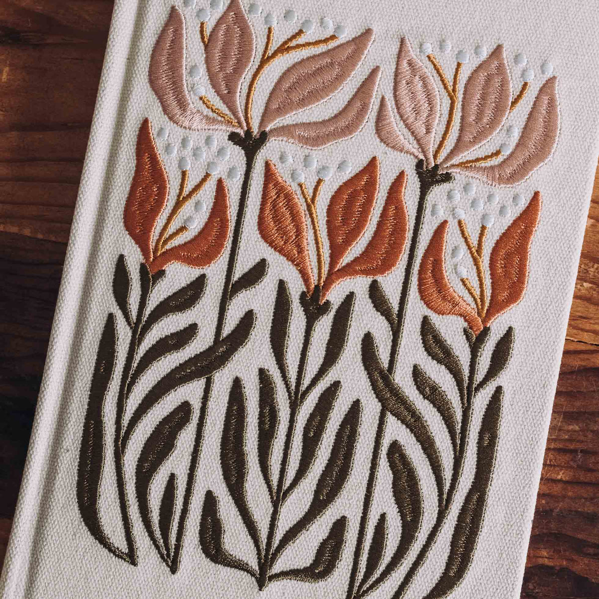 Cream hardcover journal with embroidered design of stylized lily and sunflower stems in rust, orange, and black