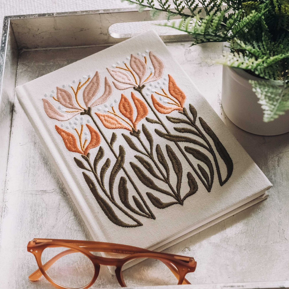 Cream hardcover journal with embroidered orange lilies fading into deep brown leaf stems, displayed with tortoiseshell glasses and succulent plant.