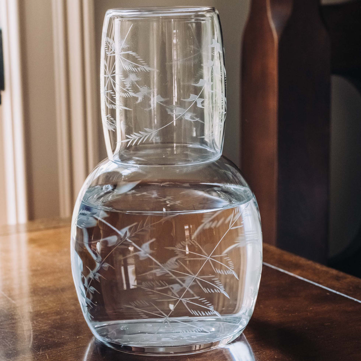 Clear hand-blown crystal carafe set with hand-engraved fern design, featuring stacked upper pouring vessel and lower serving bowl on wood table.