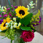 Bouquet of sunflowers, roses, and other flowers in a clear vase on a wooden surface.