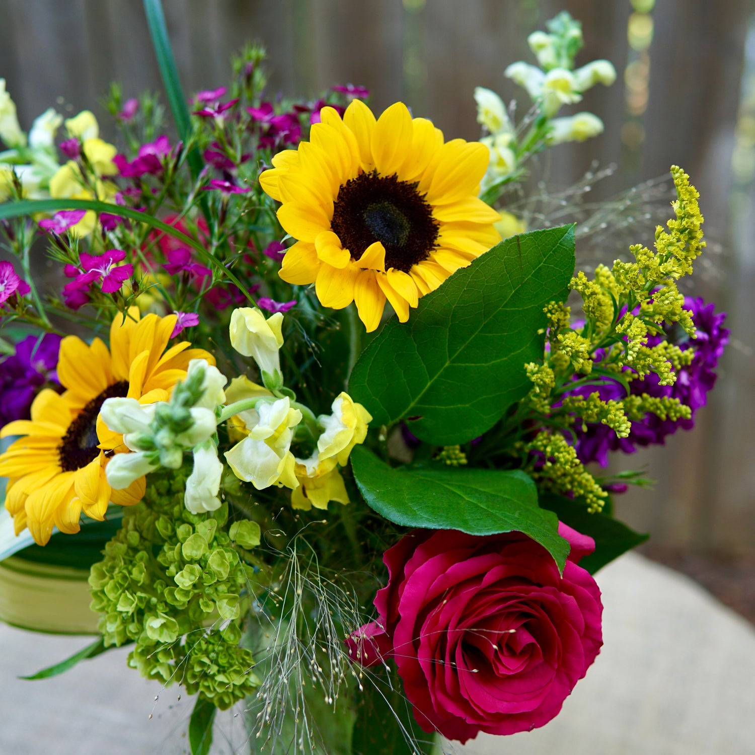 Bouquet of sunflowers, roses, and other flowers in a clear vase on a wooden surface.