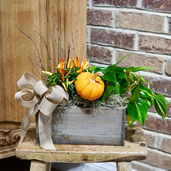 Decorative arrangement with a small pumpkin, greenery, and a bow in a wooden box on a stool against a brick wall.