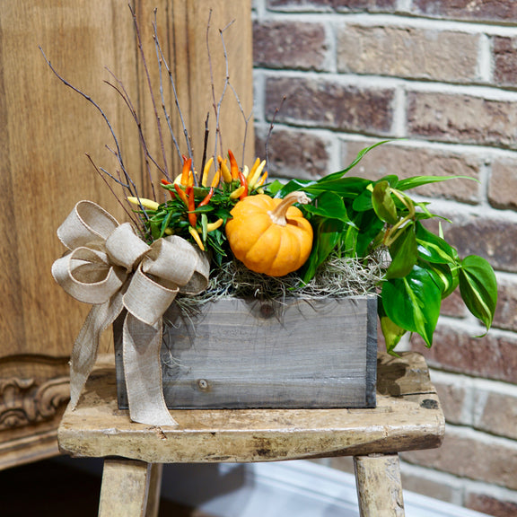 Decorative arrangement with pumpkins, greenery, and a bow on a wooden stool against a brick wall.