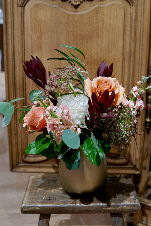 Floral arrangement in a metallic vase on a wooden stool with a wooden panel background
