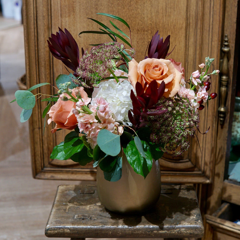 Floral arrangement in a gold vase on a wooden stool with an ornate wooden cabinet in the background.