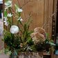 Floral arrangement with greenery and white flowers in a wooden box on a wooden chair against a wooden wall.