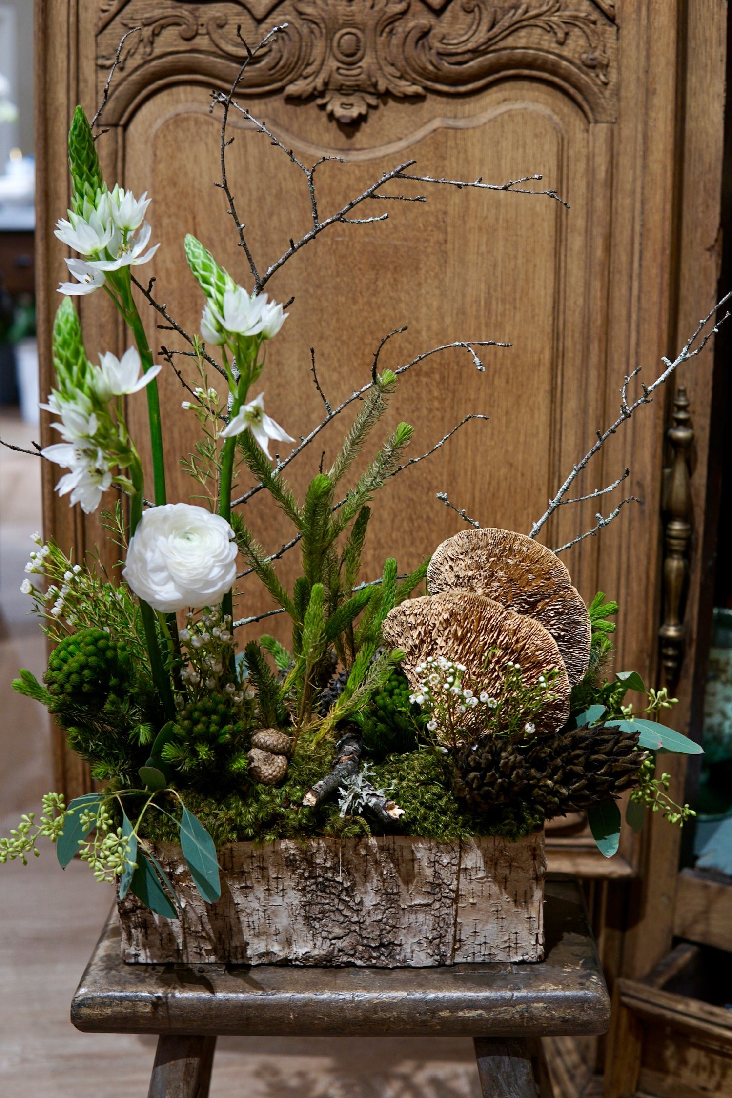 Floral arrangement with greenery and white flowers in a wooden box on a wooden chair against a wooden wall.