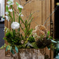 Floral arrangement in a wooden box on a rustic wooden stool with a decorative wooden panel in the background.