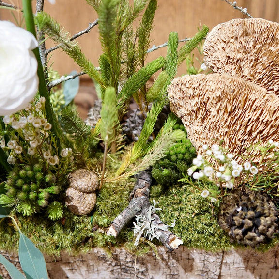 Decorative arrangement with greenery, mushrooms, and white flowers on a wooden surface.