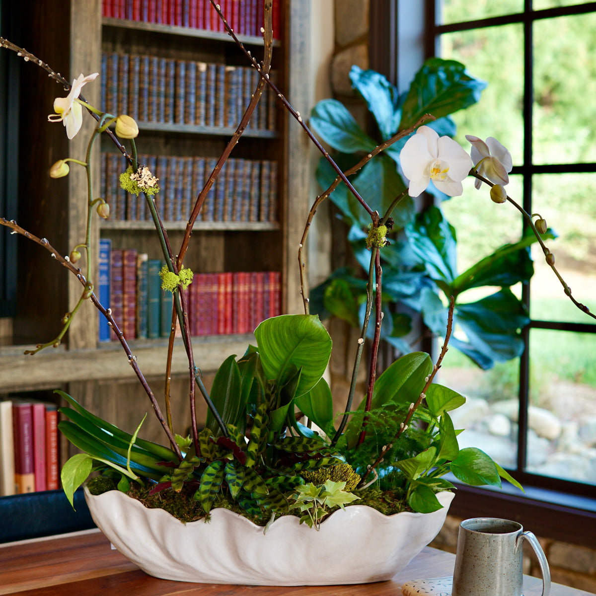 Decorative plant arrangement in a white bowl on a wooden table with books and a mug in the background.