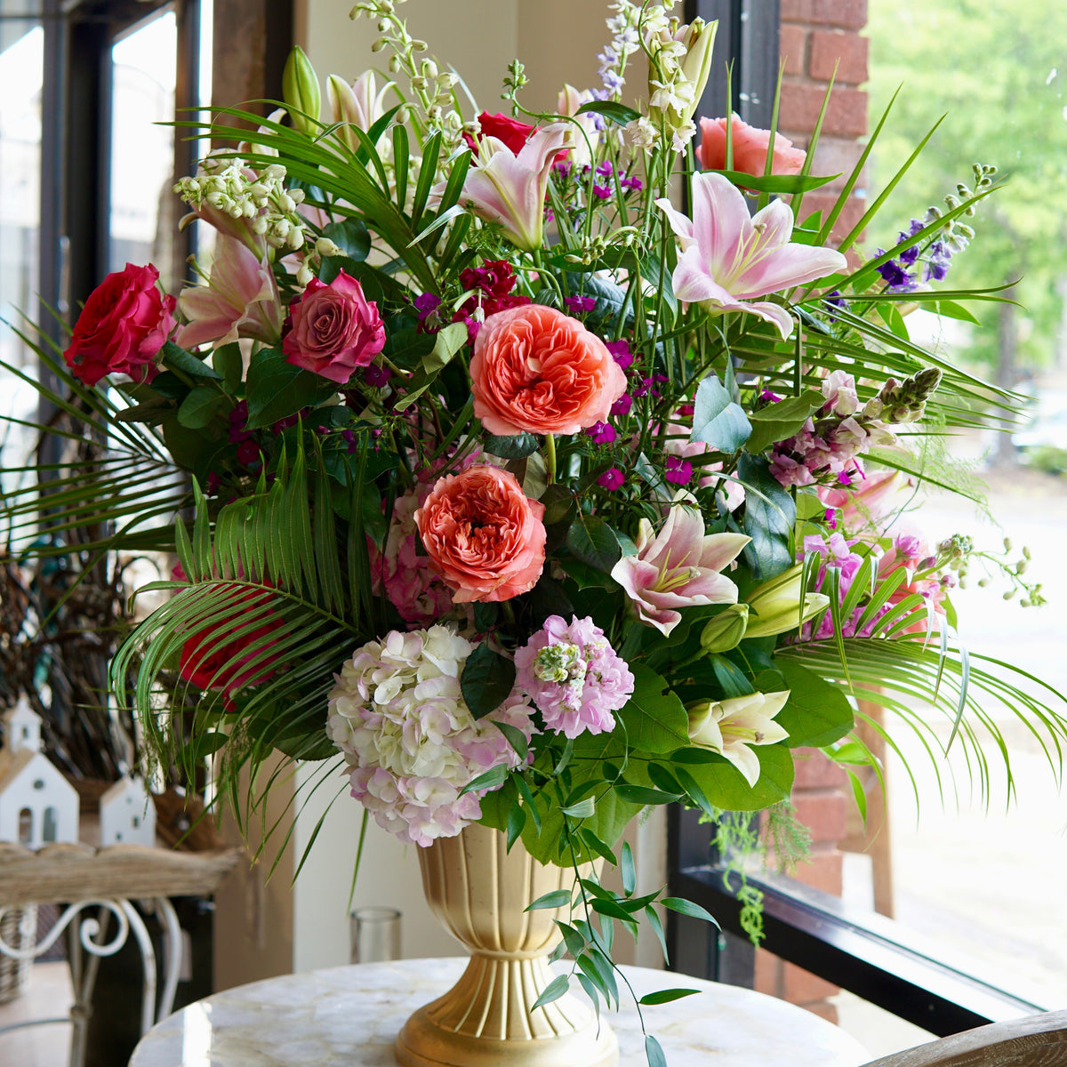Lush flower arrangement of coral and red roses, pink lilies, and cream carnations in gold pedestal vase