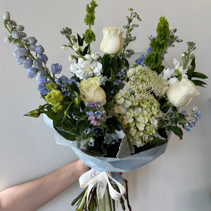 Hand-tied bouquet of cream roses, blue delphinium, white hydrangea, stock flowers, Queen Anne's lace, and green foliage, wrapped in tissue with white ribbon