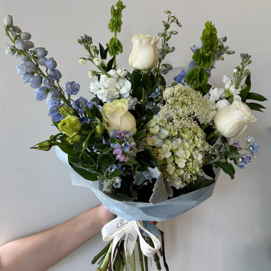 Hand-tied bouquet of cream roses, blue delphinium, white hydrangea, stock flowers, Queen Anne's lace, and green foliage, wrapped in tissue with white ribbon