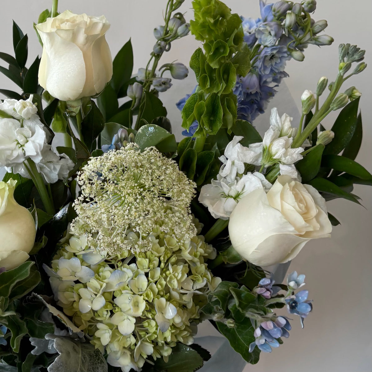 Cream roses and white hydrangeas arranged with blue delphinium, white filler flowers, and lush green foliage in a hand-tied bouquet.