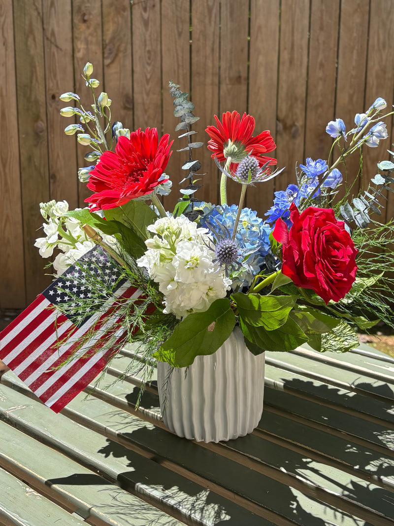 Colorful flower arrangement on a wooden deck with a chair and American flag in the background.