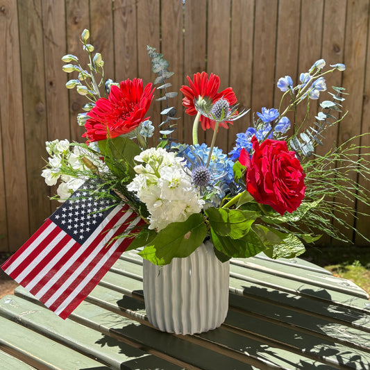 Bouquet of red, white, and blue flowers with an American flag on a wooden table outdoors.