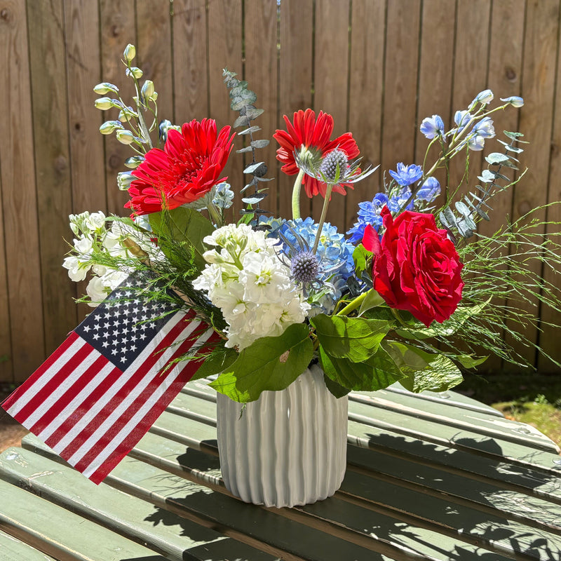 Bouquet of red, white, and blue flowers with an American flag on a wooden table outdoors.