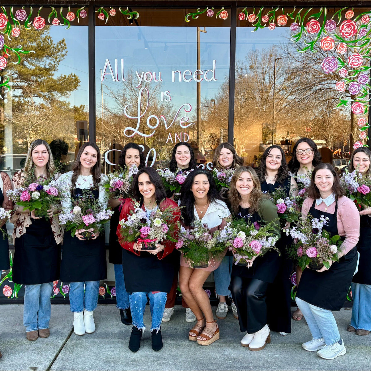Group of students holding mixed flower arrangements in pink, white, and red blooms outside storefront window with floral frame and "All you need is Love and flowers" text.