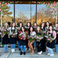 Group of students holding mixed flower arrangements in pink, white, and red blooms outside storefront window with floral frame and "All you need is Love and flowers" text.