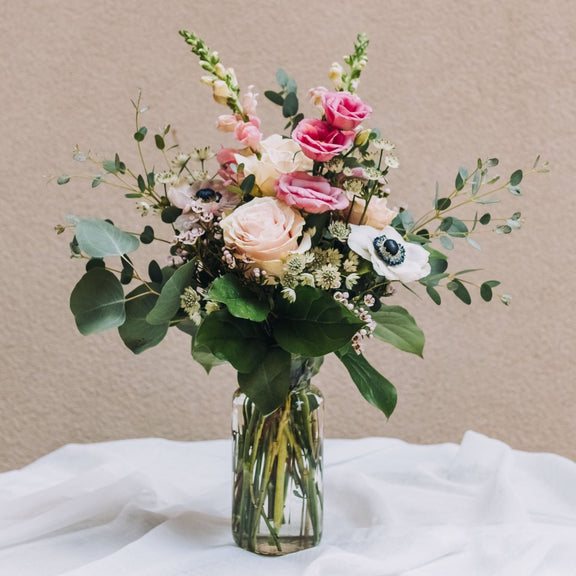 Bouquet of flowers in a clear glass vase. The bouquet is a mix of light pink and white blooms, including roses, anemones, and possibly lisianthus and snapdragons. There's also greenery, likely eucalyptus, creating a lush and romantic look. The flowers are arranged in a clear glass jar, sitting on a white fabric-covered surface against a muted beige background. The overall style is simple and elegant.