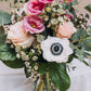 Bouquet of flowers in a clear glass vase. The bouquet is a mix of light pink and white blooms, including roses, anemones, and possibly lisianthus and snapdragons. There's also greenery, likely eucalyptus, creating a lush and romantic look. The flowers are arranged in a clear glass jar, sitting on a white fabric-covered surface against a muted beige background. The overall style is simple and elegant.