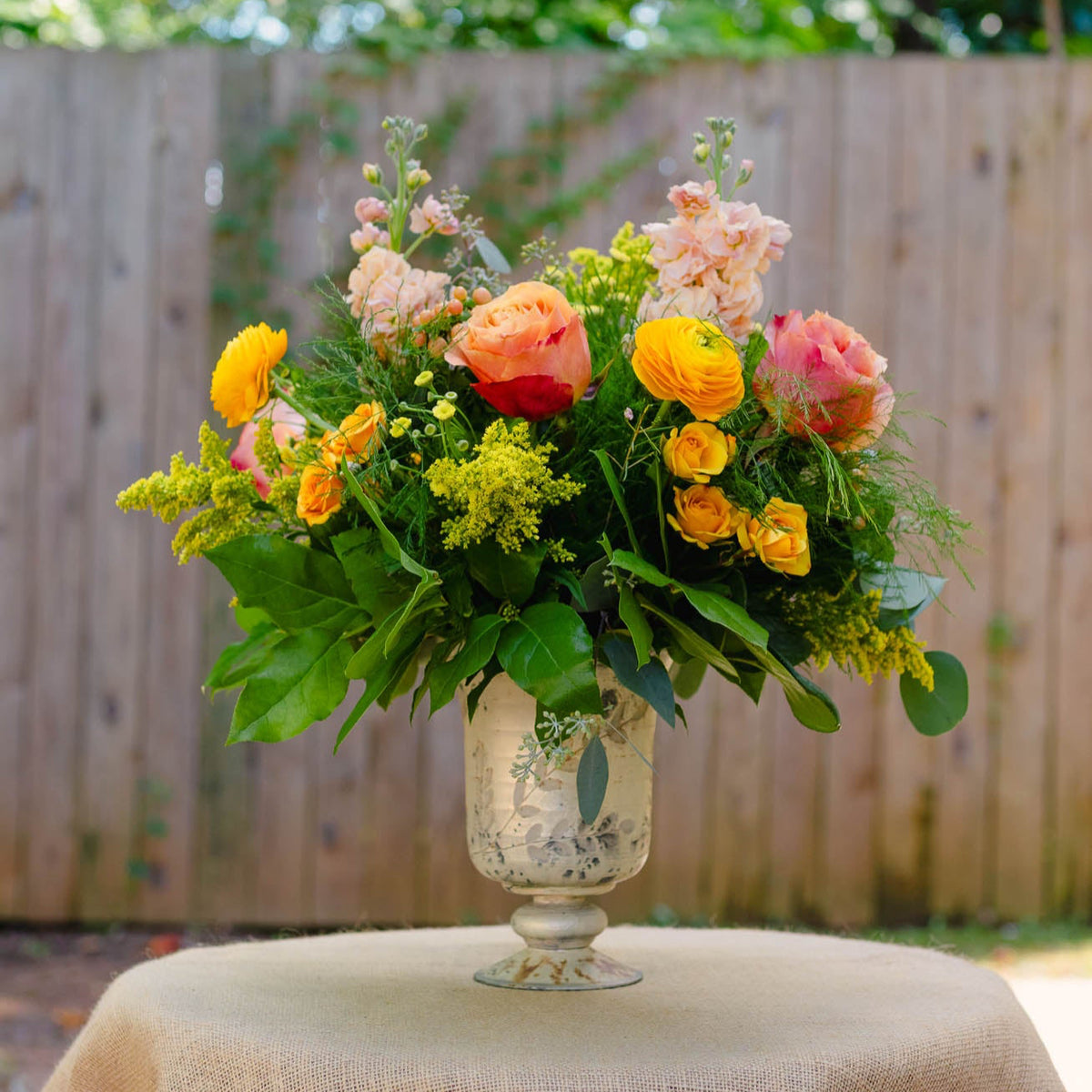 Floral arrangement in a glass vase on a table outdoors with a wooden fence in the background.