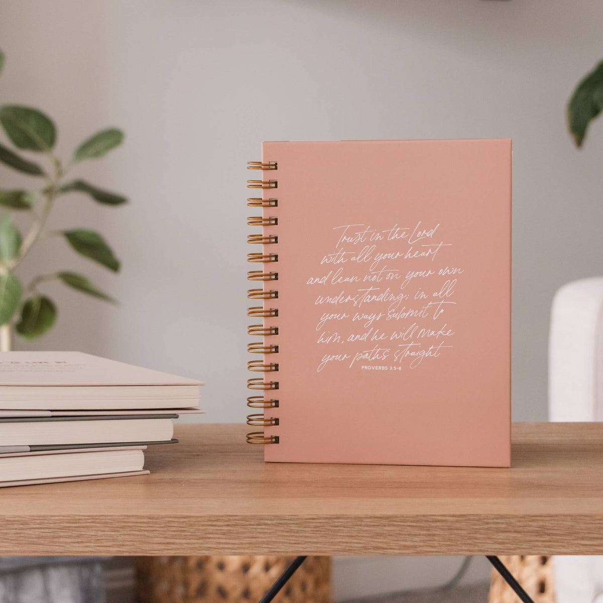 Peach hardcover spiral journal with "Trust in the Lord" verse in white script, displayed on wooden shelf with stacked books and potted plants