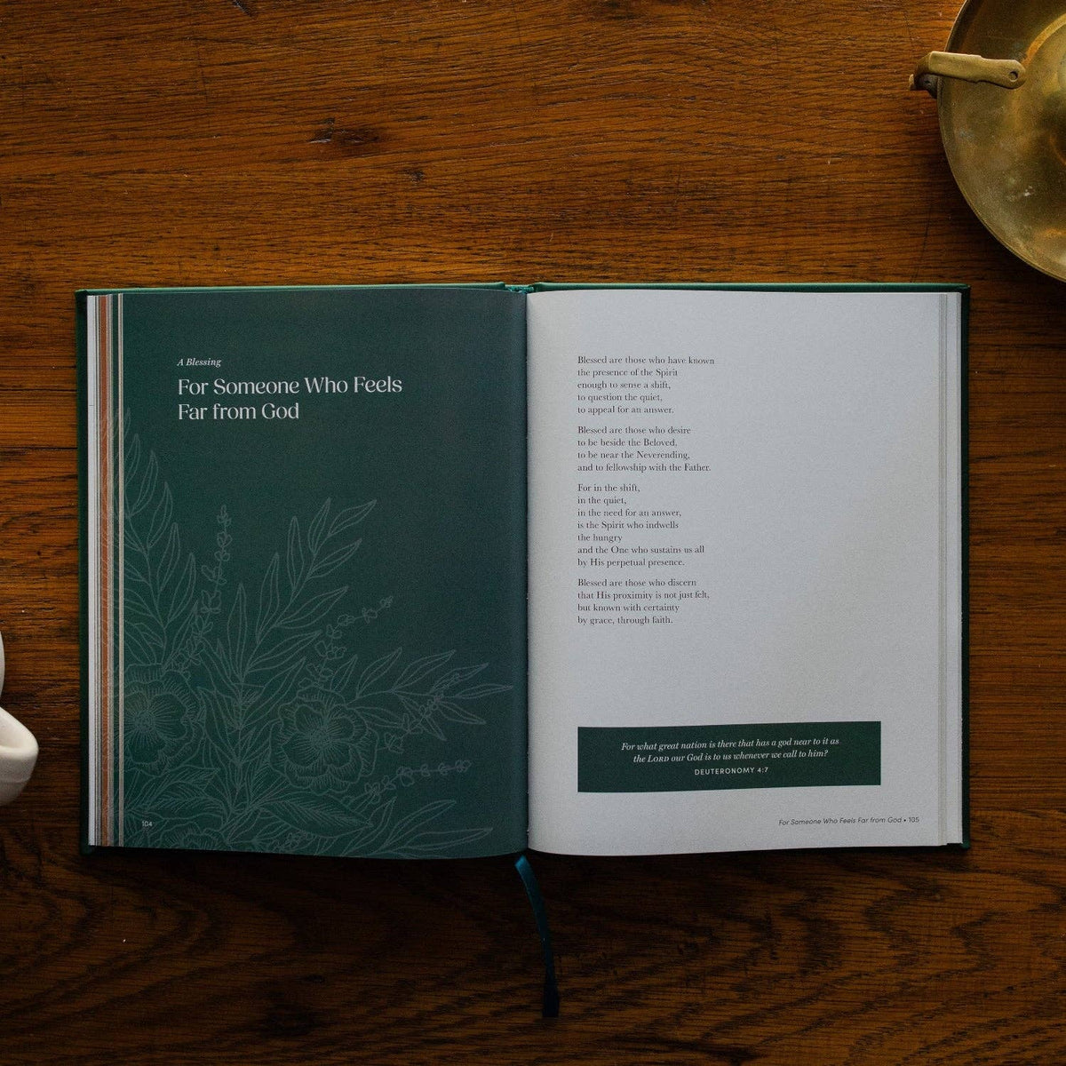 Open prayer book with forest green left page titled 'For Someone Who Feels Far from God' and botanical illustration, cream right page with prayer text on wood surface.