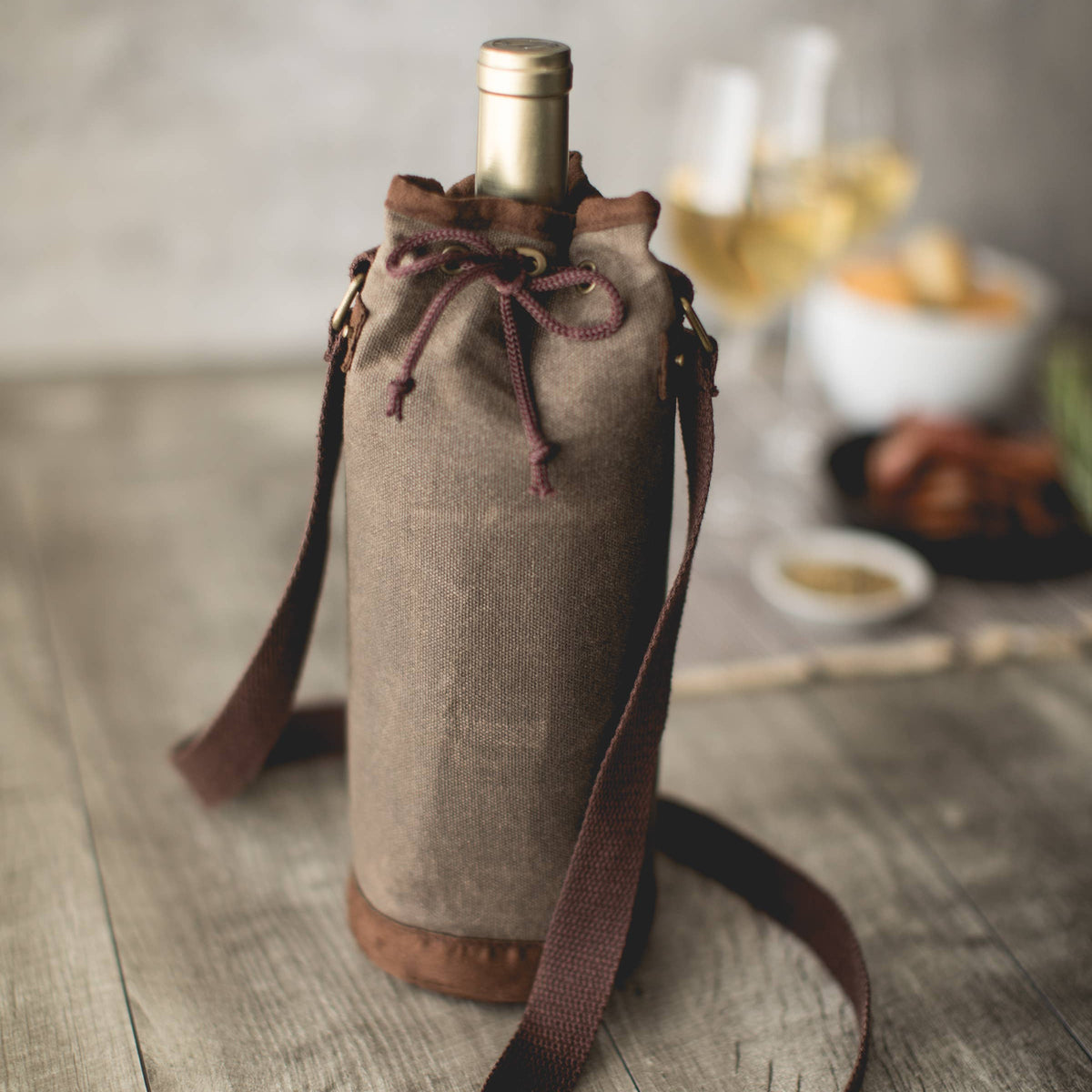 Taupe waxed canvas wine tote bag with burgundy drawstring and shoulder strap, holding wine bottle on wooden table
