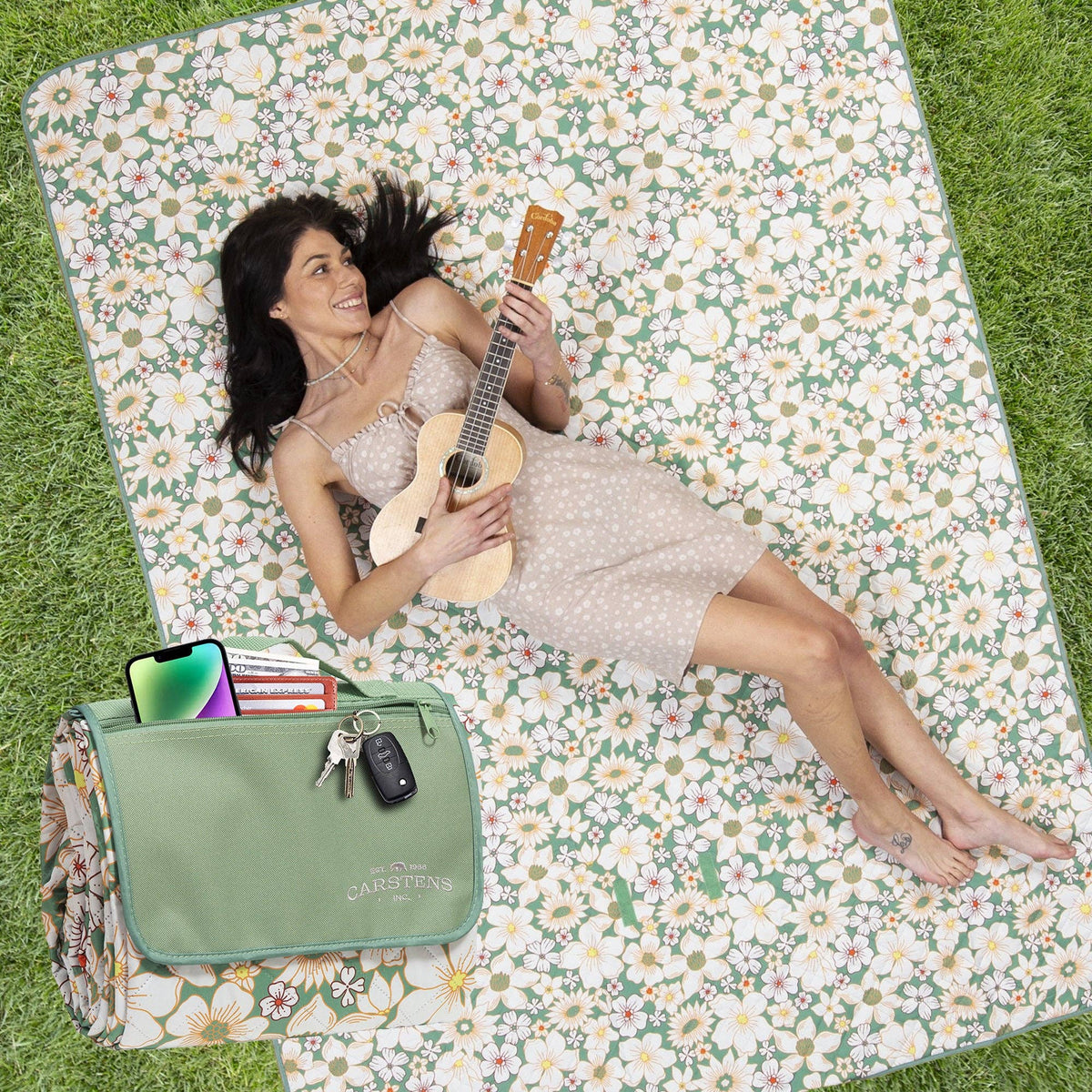 Woman reclining on cream daisy-patterned picnic blanket, holding ukulele, with green tote bag beside her on grass.