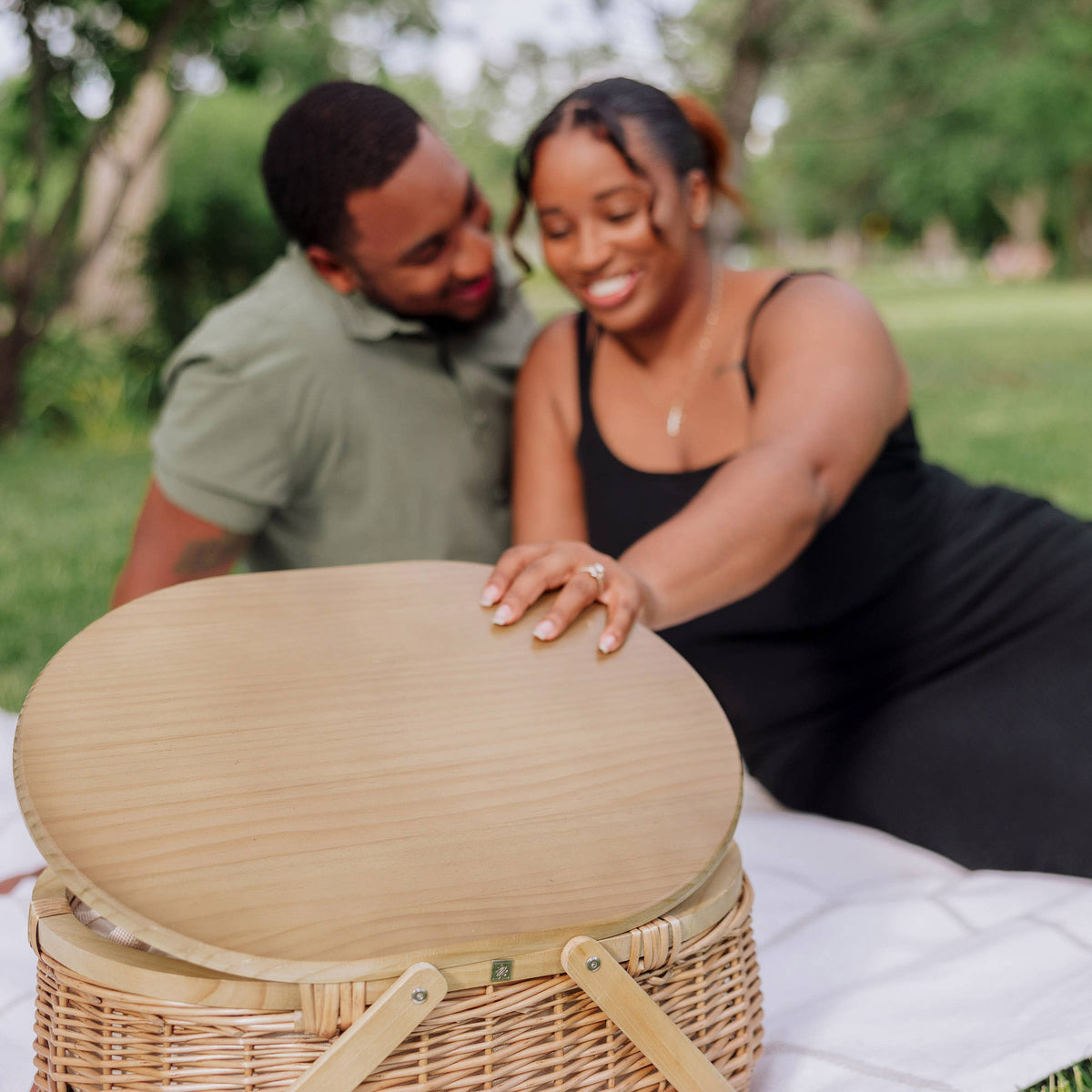 Beige wicker picnic basket with light wood oval lid on white picnic blanket in a sunny park setting.