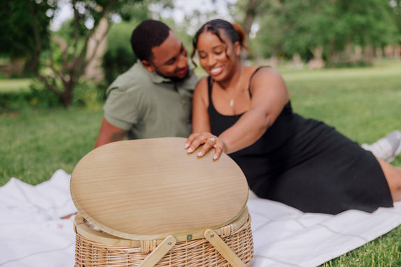Beige wicker picnic basket with light wood oval lid on white picnic blanket in a sunny park setting.